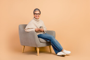 Full body photo of retired woman wear turtleneck sit armchair posting content in internet with phone isolated on beige color background