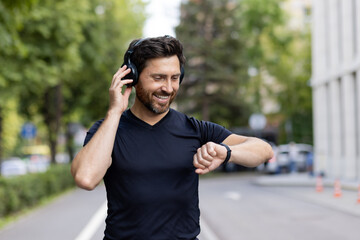 Close-up photo of a smiling young male athlete standing outside wearing headphones and looking contentedly at a smart watch on his wrist