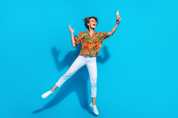 Fototapeta na wymiar Full body photo of active excited lady jumping hold smart phone take selfie arm waving isolated on blue color background