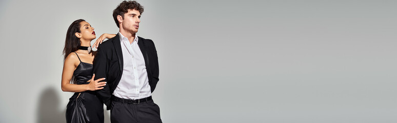 seductive young woman posing in black slip dress near handsome man in suit on grey backdrop, banner