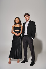handsome man in suit standing with hand in pocket near gorgeous and brunette woman on grey backdrop