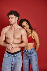 sensual young woman in bra and denim jeans leaning on shirtless man on red background, affection