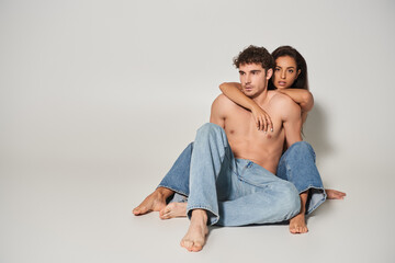 barefoot and sexy woman in blue jeans embracing her shirtless boyfriend on grey background, love