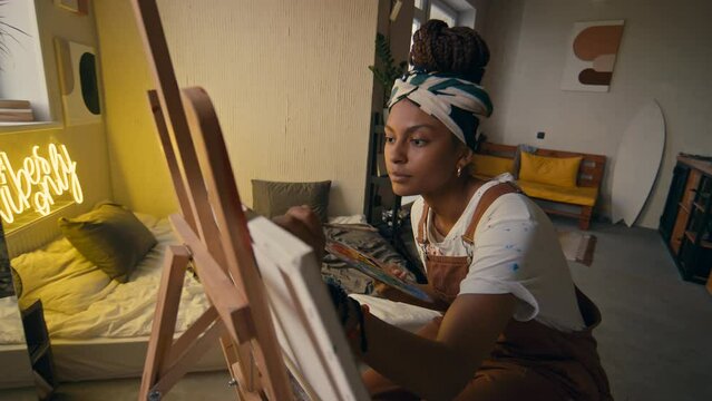 Side medium footage of female African American left-handed artist working on new painting using paintbrushes and palette in modern apartment