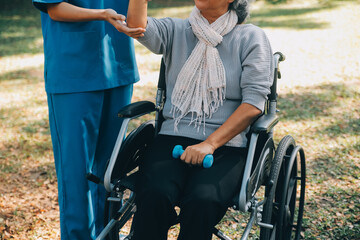 Young nurse or physiotherapist in scrubs helping a happy retired old woman do fitness exercises with light weight dumbbells at home. Concept of physiotherapy for seniors