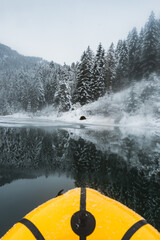 A tranquil winter scene with the front of a yellow kayak navigating a misty, snow-covered river lined by frosty trees.