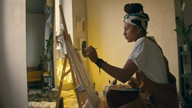 Medium side footage of young left-handed African American female artist wearing hairband and jumpsuit creating new painting using easel, canvas, paint and paintbrushes in modern studio with neon light