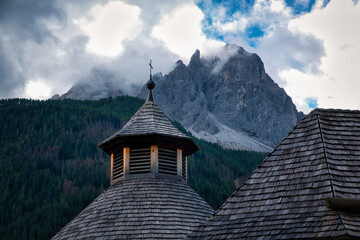 Blick auf die Spitze eines hölzernen Kirchturms mit Kreuz vor Bergpanorama mit in Wolken...