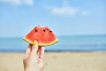 Fototapeta na wymiar A persons hand is showcased against a serene beach backdrop, holding a ripe, juicy slice of watermelon.