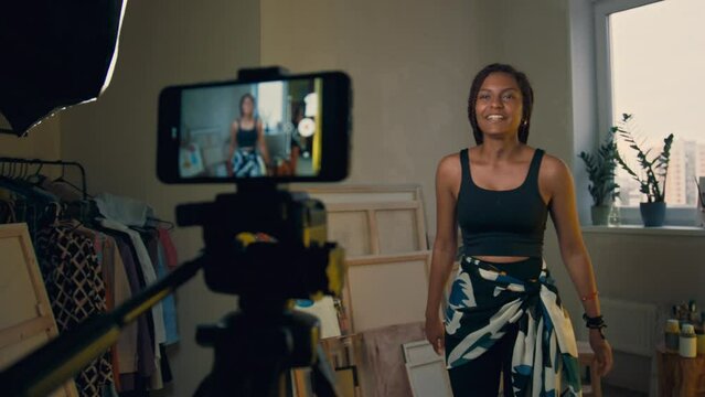 Closeup of phone on tripod filming young African American woman with box braids wearing fitness suit while talking to camera introducing herself and explaining dance moves technique at home