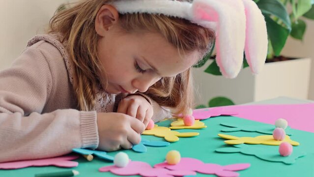 Child girl in rabbit ears creating Easter paper colored garland Easter bunny. Cute children's crafts for Easter party at home. Holiday Art Activity Kid. Fine motor skills, creativity and hobby DIY