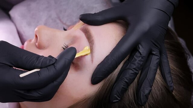The permanent makeup artist applies wax to remove excess hairs along the contour of the eyebrows. Permanent makeup procedure, performing PMU of eyebrows