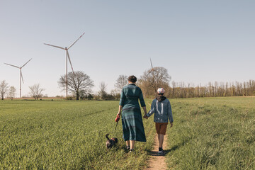 Mother, daughter and small black dog walking in green fields next to wind turbines