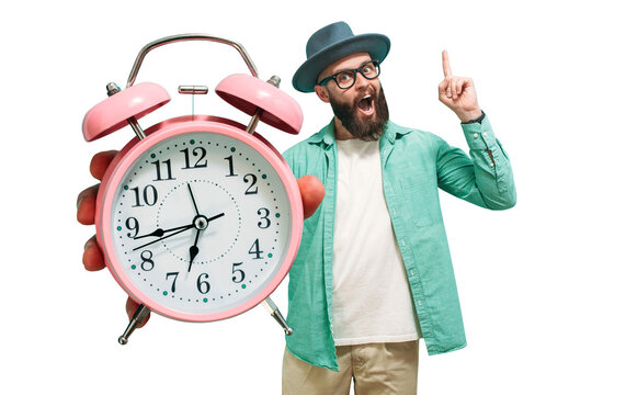 Young crazy bearded charismatic hipster holding a closeup of a huge pink alarm clock pointing at it isolated. It's time to.