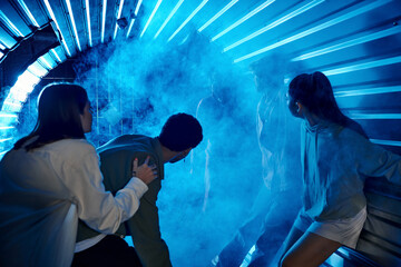 scared diverse friends standing in tunnel near freaky people in gas masks, quest room participation