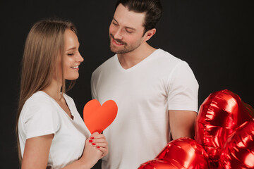 Beautiful couple isolated on grey background. Attractive young woman and handsome man are hugging and smiling with air balloons in shape of heart in hands. Happy Saint Valentine's Day!