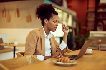 Side profile of a serious-looking African woman, working online at the cafe, using a laptop.