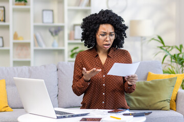 Shocked african american woman sitting on sofa at home, working with laptop and documents and...