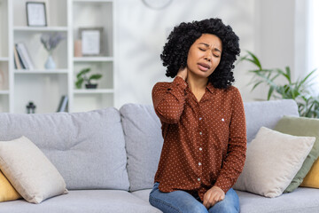 African American young woman sitting at home on sofa tired, closing eyes resting and doing neck...
