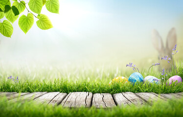Painted easter eggs in in the grass celebrating a Happy Easter in spring with a rabbit in a distant meadow with a bright sunlight background with copy space & rustic wooden bench to display products.