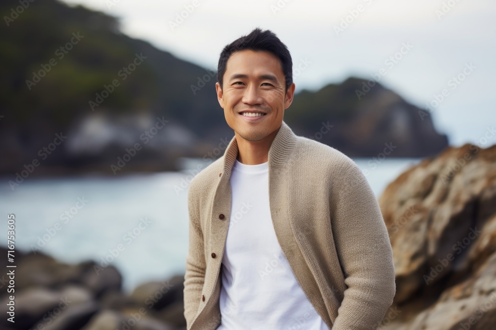 Wall mural Portrait of a satisfied asian man in his 30s wearing a chic cardigan against a rocky shoreline background. AI Generation