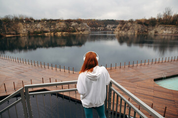 Back view of red hair sporty girl with headphones on her head standing on balcony and enjoy view of beautiful lake with wooden pier Zakrzowek Quarry, Krakow