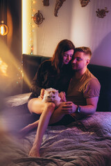 A couple of a guy and a girl are sitting on a bed in a room with colored lighting. The guy caresses...