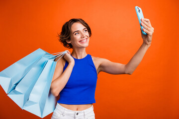 Photo of gorgeous positive person hold store bags smart phone make selfie isolated on orange color background
