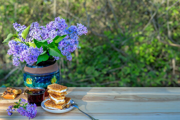 Teatime in spring garden on sunny evening. Tea with Viennese waffles under bouquet of lilacs. Still life with sweets in rustic style. Copy space, text space, postcard.