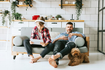 family dad young man with phone and son teenage cute boy with tablet and headphones sit on couch in...