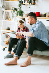 family dad young man with phone and son teenage cute boy with tablet and headphones sit on couch in cosy apartment with pet poodle dog, quality time with your family, gadget addicted