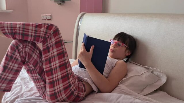 Beautiful girl child reads interesting stories before bed. Young teenager with glasses holding a hardback book lying in bed