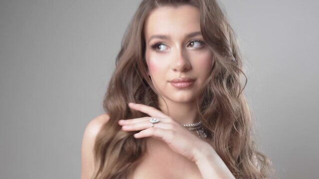 A glamorous model girl with evening makeup and hairstyle shows off an expensive beautiful necklace with diamonds and pearls around her neck and a ring on her finger, posing in the studio.