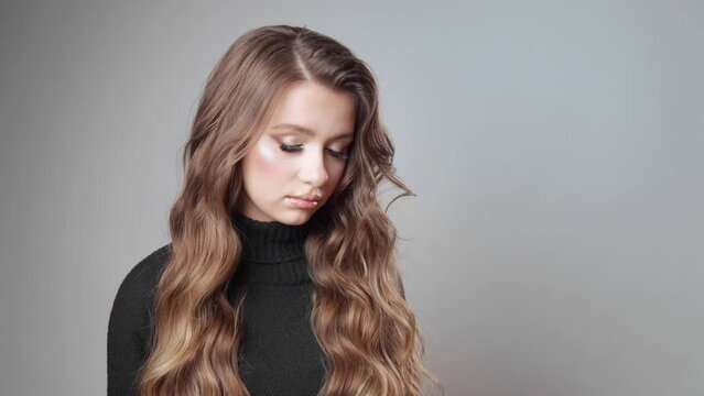 A sad, upset girl with long brown hair in a bad mood, dressed in a warm black sweater, looks at the camera with disappointment, reproach and resentment in her eyes. A model poses in the studio.