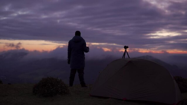 A wild camping photographer drinking coffee and looking out over majestic clouds next to his camera and tripod