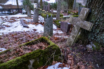 Broken old damaged cross gravestone leaning against headstone covered in snow falling and settling on ground in churchyard