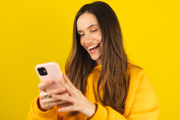 Great application or website. Portrait of excited woman using her mobile phone, isolated on yellow studio background, copy space. Positive lady holding gadget, looking at phone, texting, messaging.