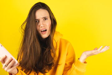 Mad and shock young brunette beautiful woman using mobile phone isolated on yellow background....
