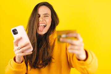 Online shopping. Portrait of smiling brunette hair woman paying with plastic credit card on smartphone app, standing with mobile phone and bank card against yellow background. Happy girl show tongue.