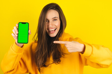 Young developer woman hold smartphone with green screen chroma key mock up recommend good application promotional sale offer, point to phone screen. Brunette girl isolated on yellow studio background.