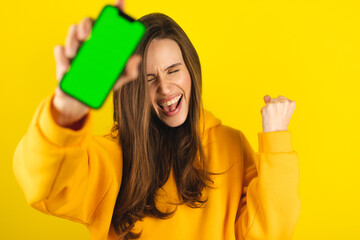 Fun woman hold in hand, point on phone green screen doing winner gesture clenching fists isolated on yellow background. Girl using smartphone apps winning online celebrating discount gift voucher.