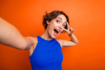 Photo of impressed excited girl dressed blue top showing v-sign cover eye tacking selfie isolated orange color background