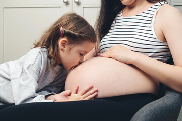 Pregnant woman with her little daughter at home. Cute little girl kisses her mother's tummy. Happy pregnant woman her child, expectation
