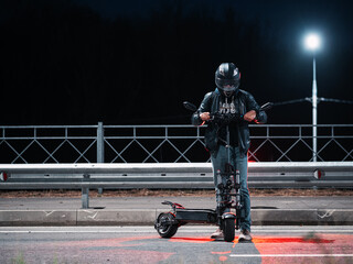 A man in a helmet poses on an electric scooter on a night road. Modern stylish and powerful urban transport.