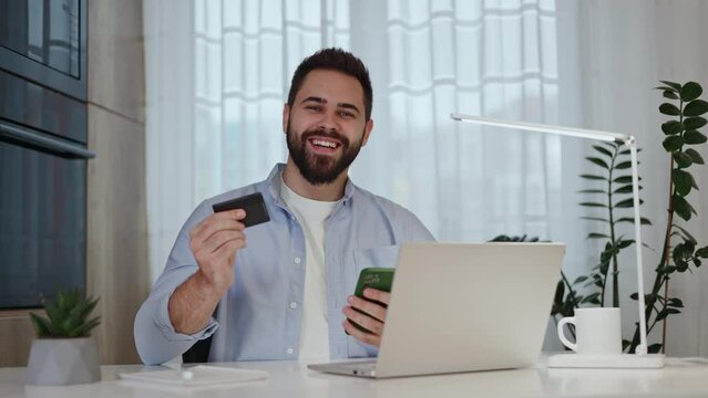 Excited male using credit card and cellphone while sitting by domestic desktop with computer. Fortunate guy receiving approval for online loan and getting notification about having money on account.