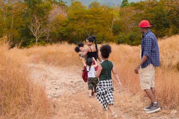 Fototapeta premium Happy African American family with adorable children weekend vacation hiking outdoors in countryside, father and mother carry children walking trail in forest park with fun, bonding relationship