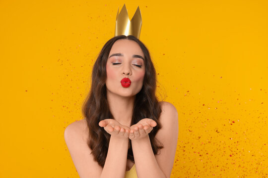Beautiful young woman with princess crown blowing kiss under falling confetti on yellow background