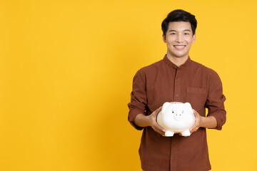 Portrait of Happy Asian man holding white piggy bank isolated on yellow background, Saving and...