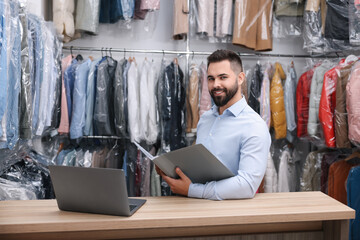 Dry-cleaning service. Happy worker with folder and laptop at counter indoors
