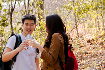 Naklejka premium Asian lovers couple male and female standing resting thirsty drinking water from plastic bottles together in summer sunny day, hiker backpack travelers happy walking hiking together outdoors activity